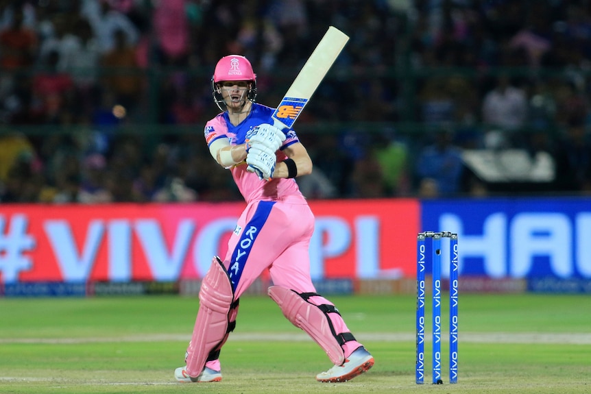 Steve Smith holds his bat up and looks behind him wearing pink cricket clothes.