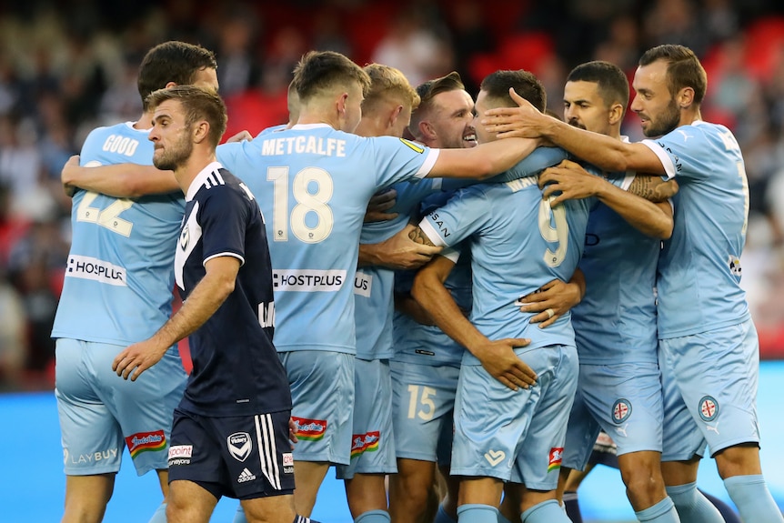 Melbourne City players celebrate in front of a Melbourne Victory player who looks disappointed