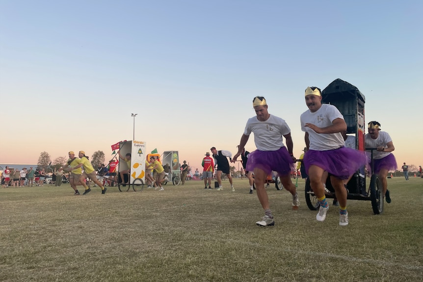 Two men in gold crowns and purple skirts running, dragging a toilet and outhouse on wheels behind them.