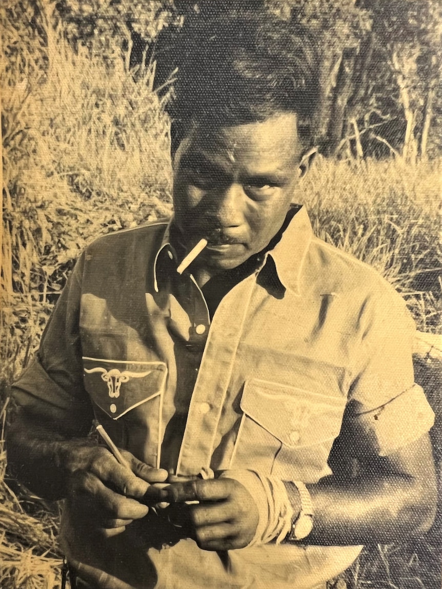 A man wearing a cowboy shirt stands in the bush with a cigarette in his mouth.  With a quiff hairstyle, he gazes into the camera
