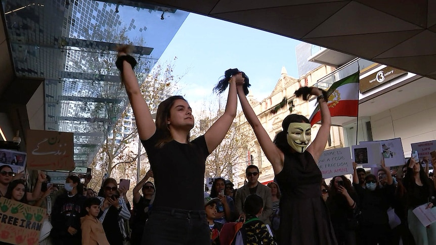 Two women hold their clumps of hair up victoriously, one wears a mask, protestors holding signs gather behind them