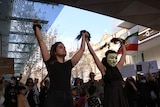 Two women hold their clumps of hair up victoriously, one wears a mask, protestors holding signs gather behind them