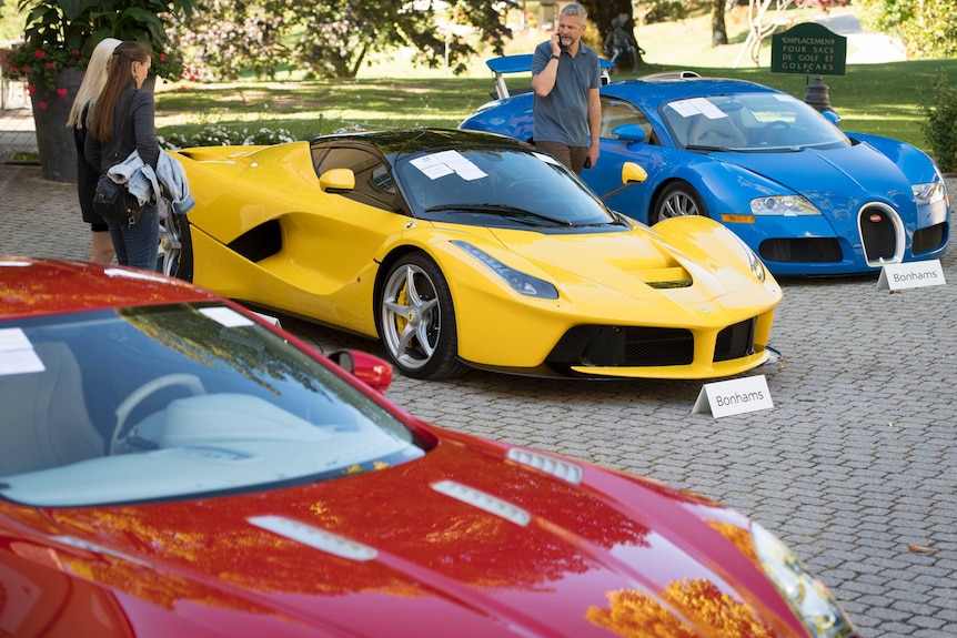 Three people are seen looking at a yellow Ferrari LaFerrari with a red Aston Martin to the left and a blue Bugatti to the right.