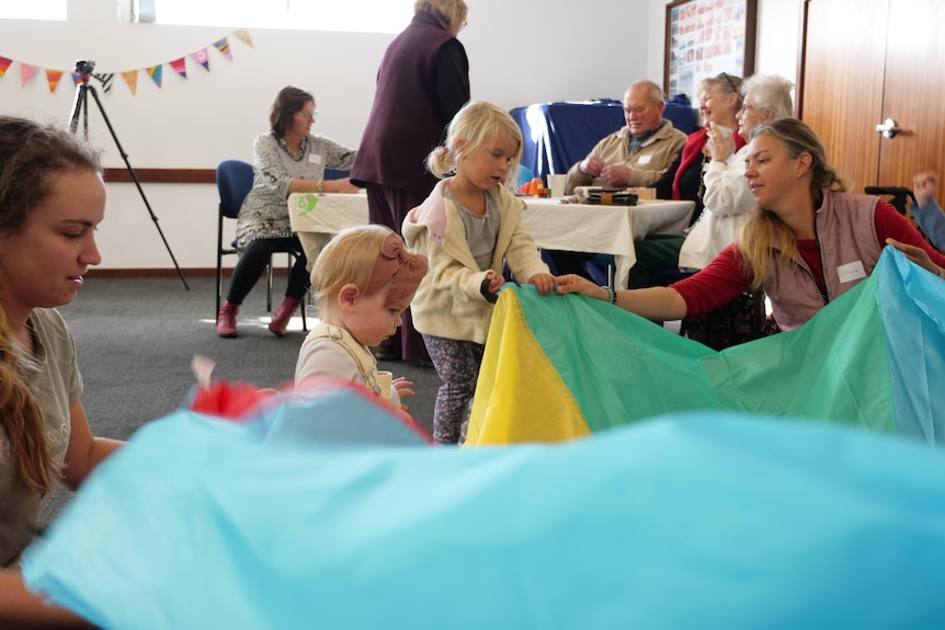 Kids lifting a bright colourful sheet with toys in the middle while people watch in the background.