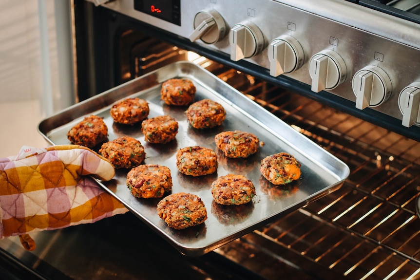 A hand in an oven mitt holds a baking tray lined with small orange patties by an open oven 