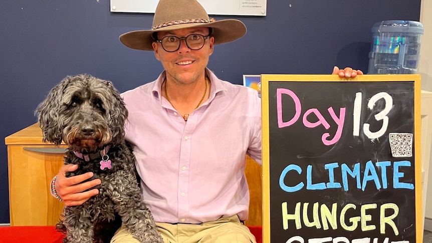 man and a dog sit on a couch with a board that reads 13 days climate hunger strike 