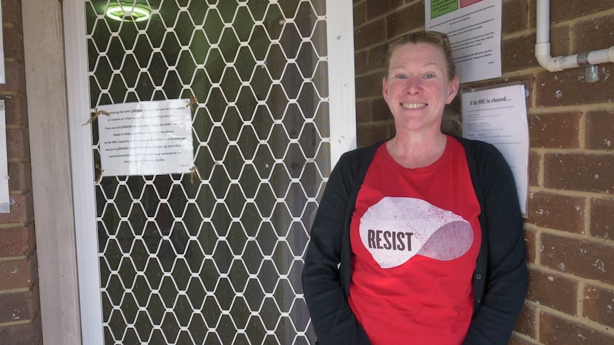 Smiling woman wearing a t-shirt with the word 'Resist' stands in the doorway of a brick building