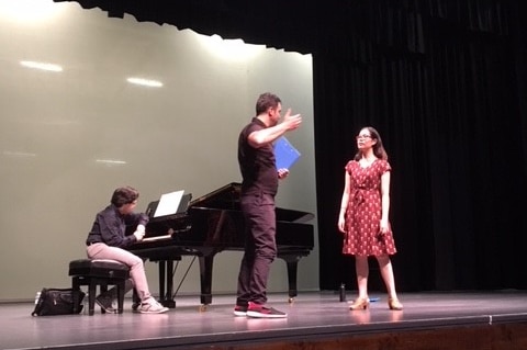 Gary stands on stage directing a participant in front of a piano