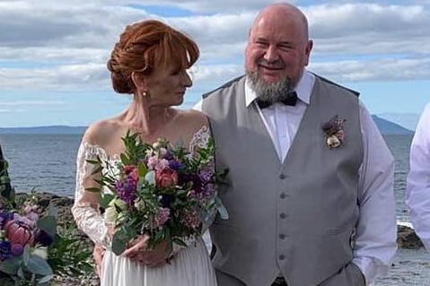 A woman with red up-do in a wedding dress holds bouqet of flowers, stands next to bald husband with beard and grey waistcoat.