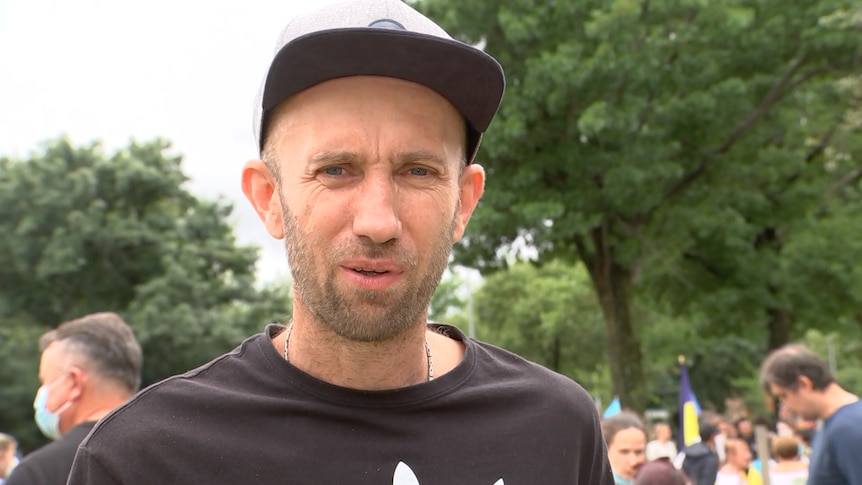 A man in a t-shirt and a cap stares at the camera, a protest going on behind him