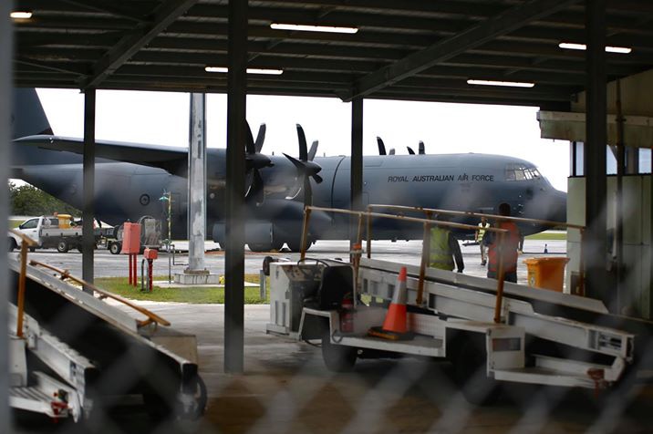 A Royal Australian Air Force Hercules C130 on the tarmac at Christmas Island's airport with airport vehicles under a hanger.