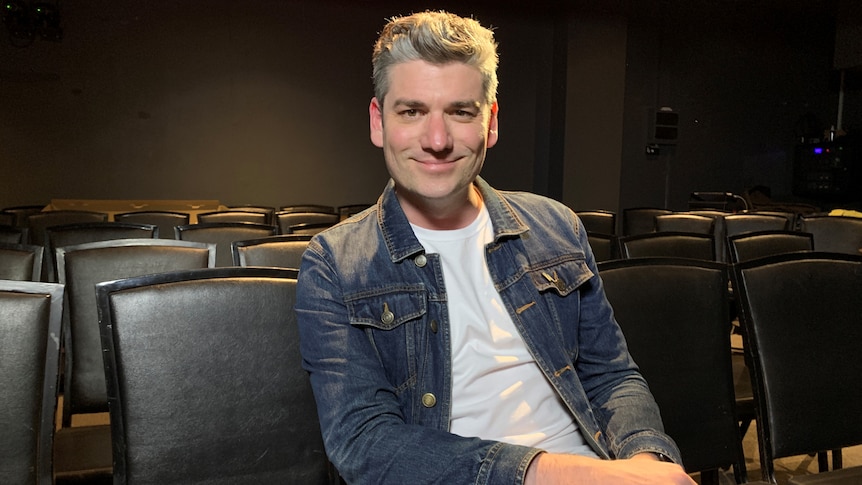 Man in his early thirties sits in a theatre wearing a white tee and denim jacket and smiles.