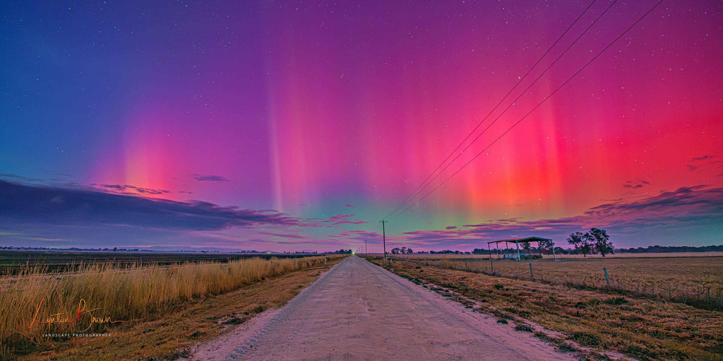 a scene of a road and grass on two sides with the sky lit up with a wide array of colourful rays.