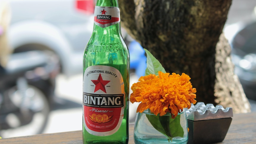 A Bintang beer bottle next to a bowl of orange flowers on a table.