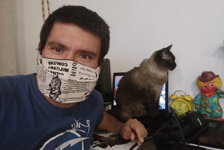 A man seated at a desk wearing a face mask with a cat next to him.