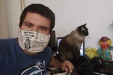 A man seated at a desk wearing a face mask with a cat next to him.