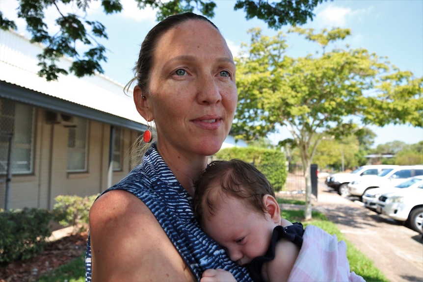 Mum with her hair pulled back wearing orange earrings cuddles a baby to her chest.