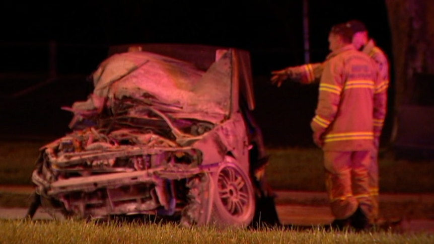 Two firemen stand next to a car on the side of the road with the engine exposed and bumper ripped off.