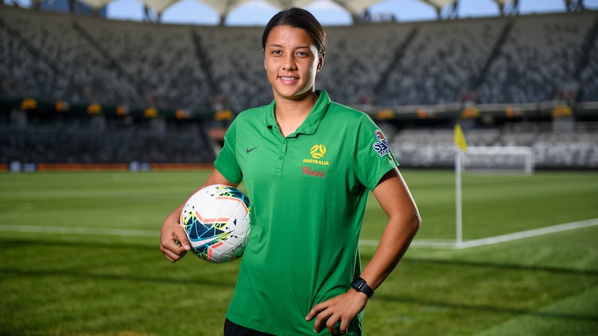 A footballer stands in a stadium holding a ball, with the corner flag behind her.