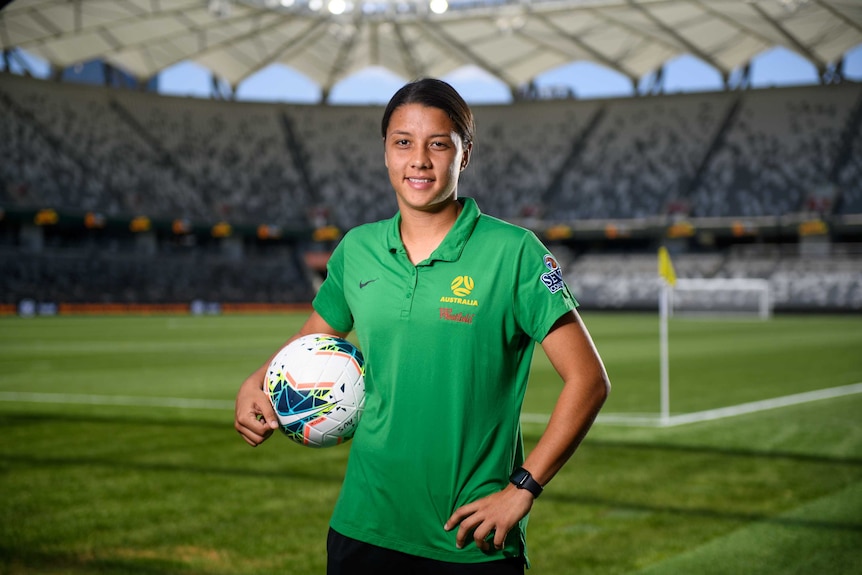 A footballer stands in a stadium holding a ball, with the corner flag behind her.