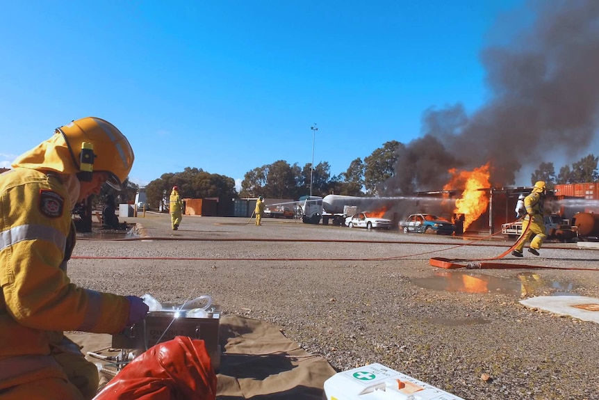 Firefighters with hoses run towards a pretend service station that is on fire.