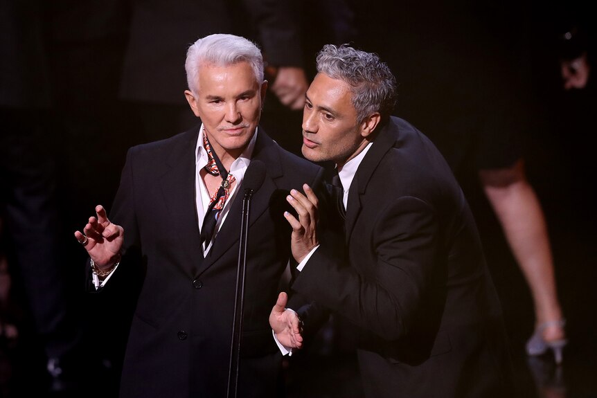 White man with combed white hair and black suit next to brown man with grey hair and black suit present award on stage.