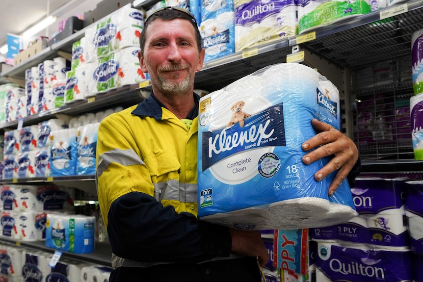 A man in a high vis shirt holds a pack of toilet paper in a supermarket aisle