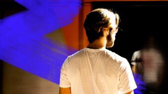 Young bearded man stands on train station platform, with a blue lightning bolt shape in the background.