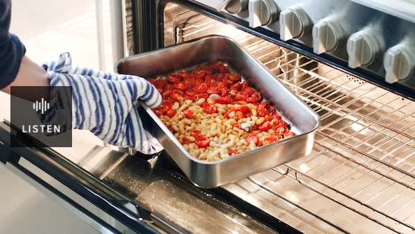 An oven mitt-clad hand puts a baking tray with cherry tomatoes, beans and garlic cloves into a large stainless steel oven. Has Audio.