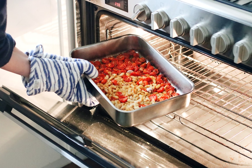 An oven mitt-clad hand puts a baking tray with cherry tomatoes, beans and garlic cloves into a large stainless steel oven.