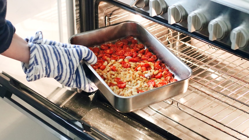 An oven mitt-clad hand puts a baking tray with cherry tomatoes, beans and garlic cloves into a large stainless steel oven.