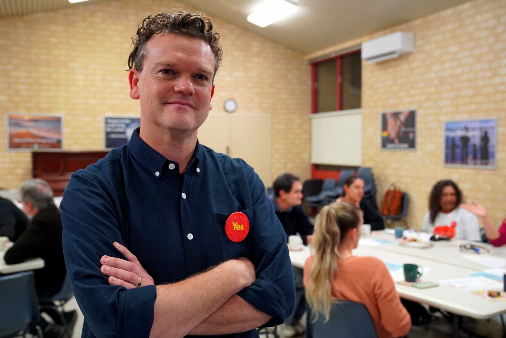 A man wearing a dark shirt with a red Yes badge on his chest stands posing with his arms folded in front of people at a table.