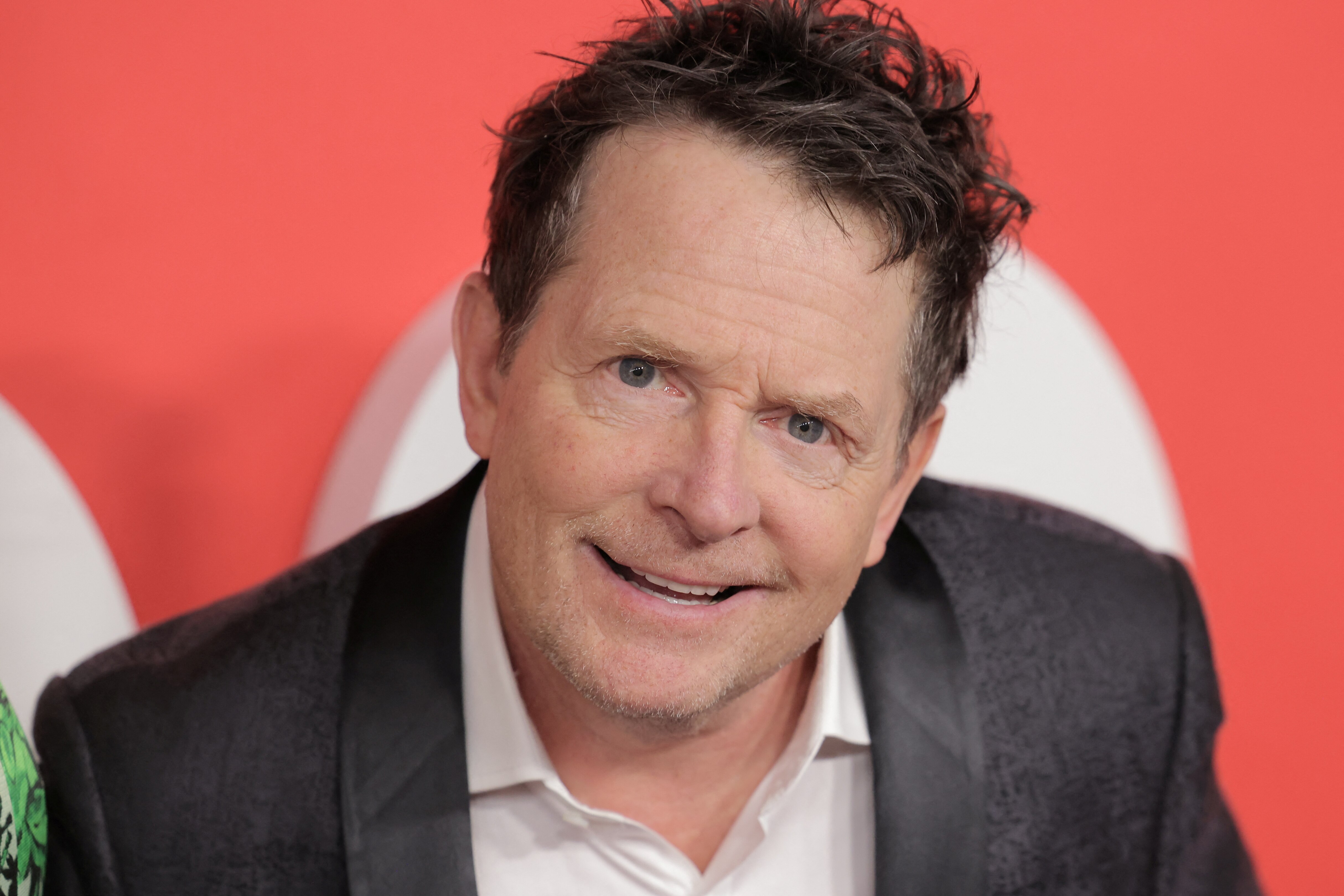 A close up of Michael J. Fox  smiling, wearing a black jacket and white button-up shirt
