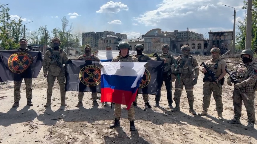 Man in military dress holds up Russian flag in fron of a row of soldiers standing behind.