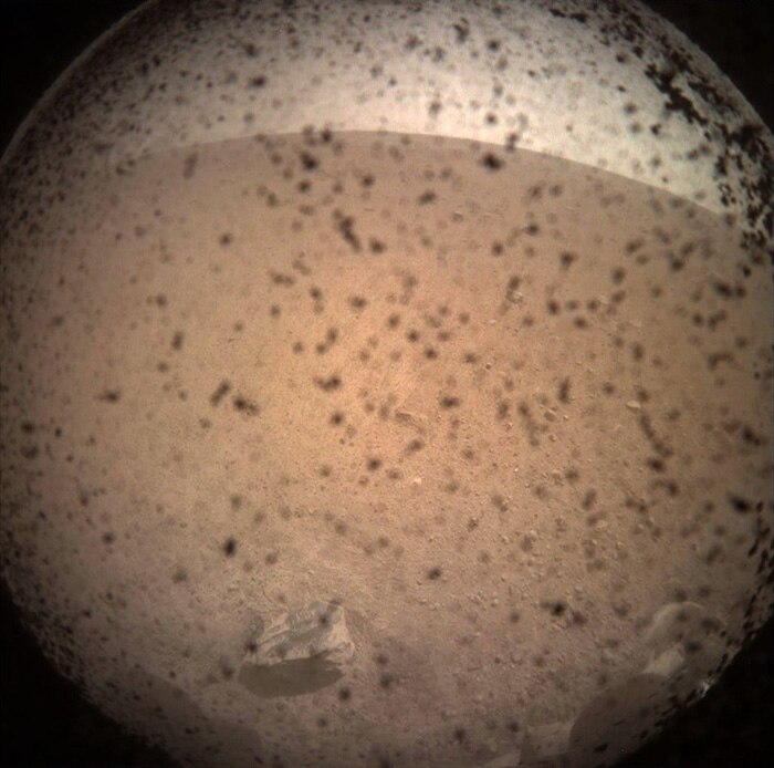 A fisheye lens photo of the red Martian surface with specks of dirt covering the camera.