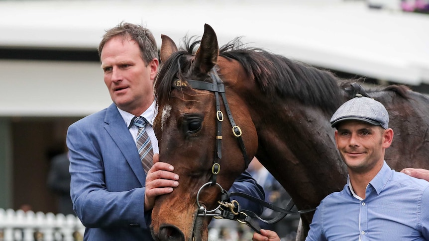 A man in a suit pats a racing horse. To their right is a jockey.