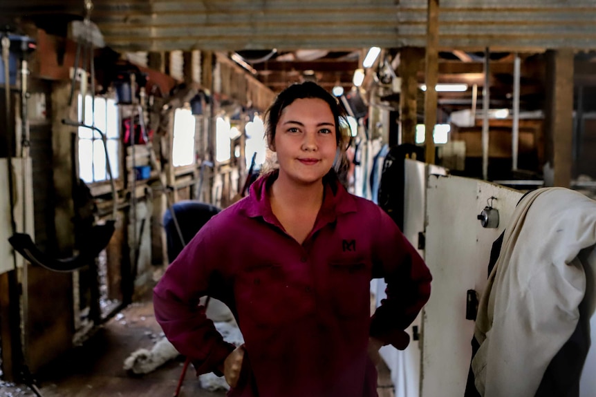 Young woman with brown hair sands with arms on side wearing purple shirt inside wooden shearing shed