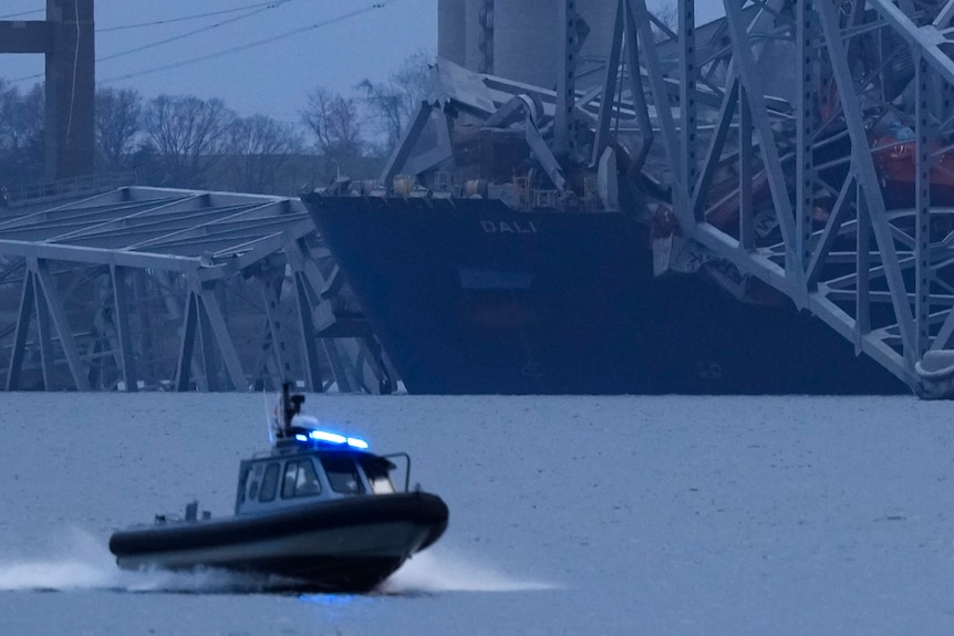 A ship with shipping containers sits with the wreckage of a bridge sitting on top of it. A police boat moves past it