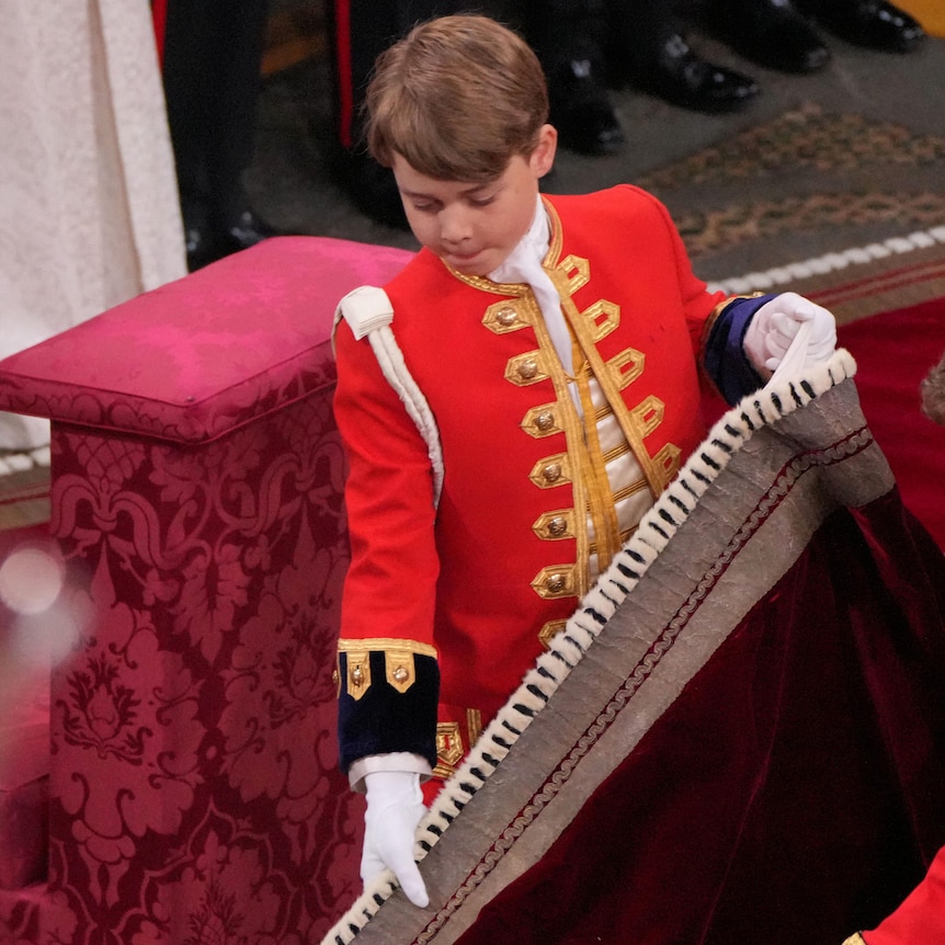 Prince George holds the train of King Charles's robe with two hands.
