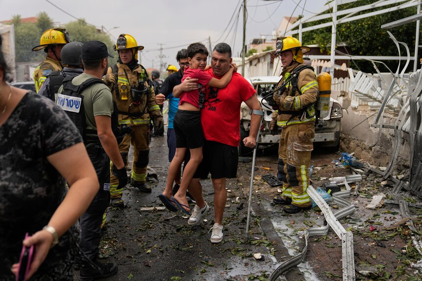 A man carries a young boy down the street amidst emergency services personnel and rubble. 