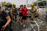 A man carries a young boy down the street amidst emergency services personnel and rubble. 