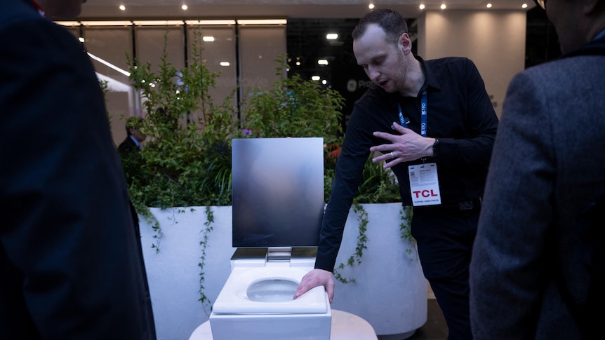 A man holds onto the seat of a smart toilet with one hand and gestures with the other as he speaks to people at a tech show