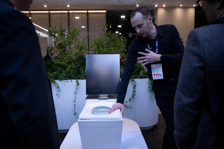 A man holds onto the seat of a smart toilet with one hand and gestures with the other as he speaks to people at a tech show