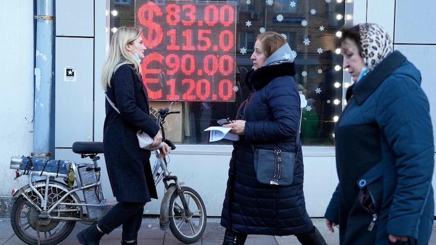 Women in Russia walk  past a screen with images showing exchange rates for currencies
