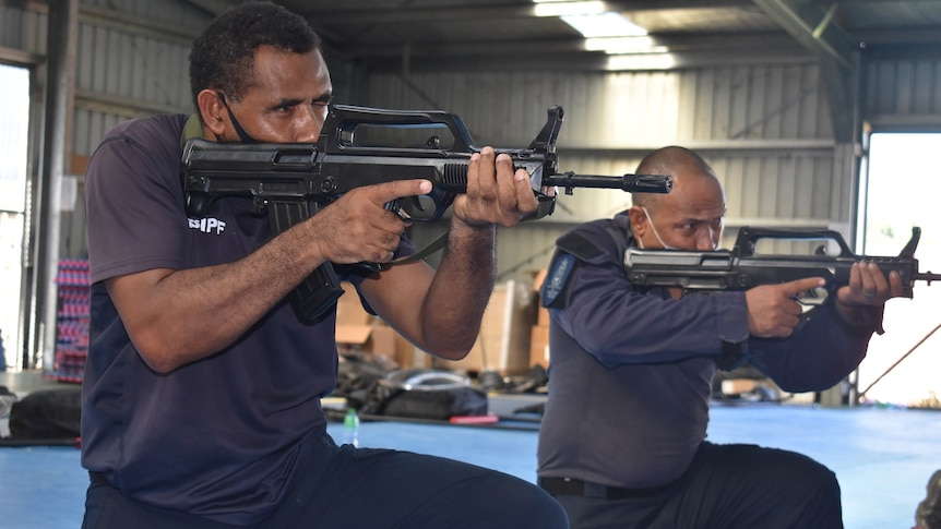 Men kneel down while aiming black semi-automatic firearms in a corrugated iron shed.