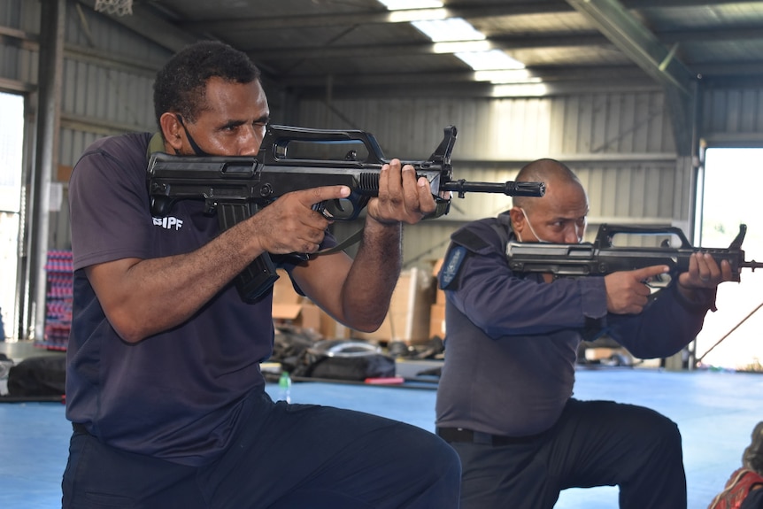 Men kneel down while aiming black semi-automatic firearms in a corrugated iron shed.