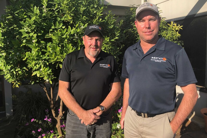 Two men stand facing the camera wearing polo shirts and caps with foliage behind them.