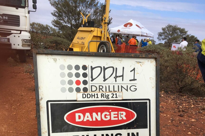 A metal drilling in progress sign on red dirt in front of a truck and large mining drill