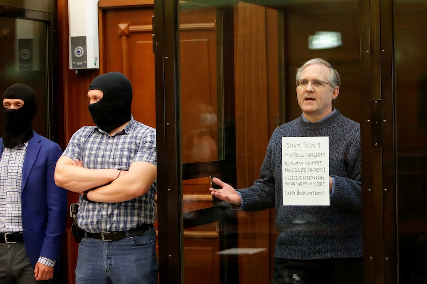 Paul Whelan stands inside a glass case. Outside the cage two men stand wearing balaclavas.
