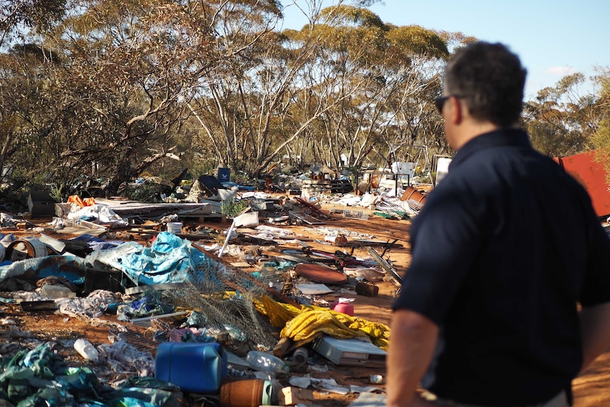 A farmer looks over an area of scrub strewn with piles of rubbish, including tarp, nappies, bottles, drums.